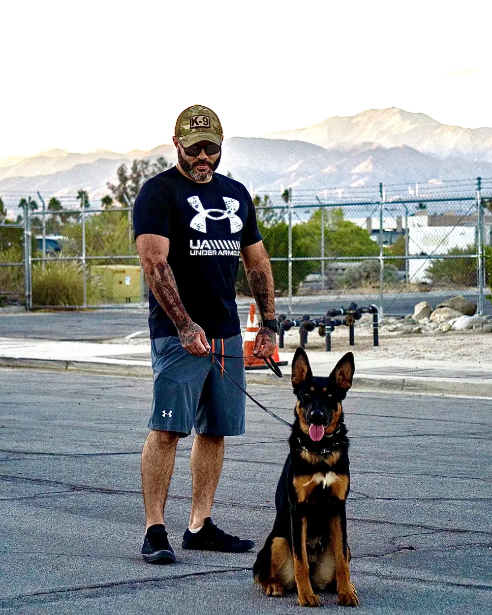 A trainer with a German Shepherd sitting at heel in an open lot, mountains behind